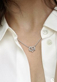 Srebrna veriga z obeskom v obliki neskončne srčne oblike, okrašena s prozornimi dragulji, na belem ovratniku srajce.