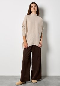 Suéter de punto acanalado beige con cuello alto y corte oversize, combinado con pantalones holgados marrones y calzado beige, sobre un fondo liso.