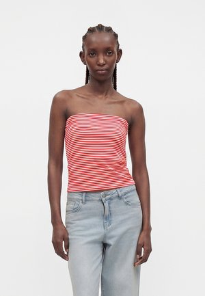 Femme aux cheveux tressés portant un haut bandeau rayé rouge et blanc et un jean bleu clair, debout devant un fond blanc uni.