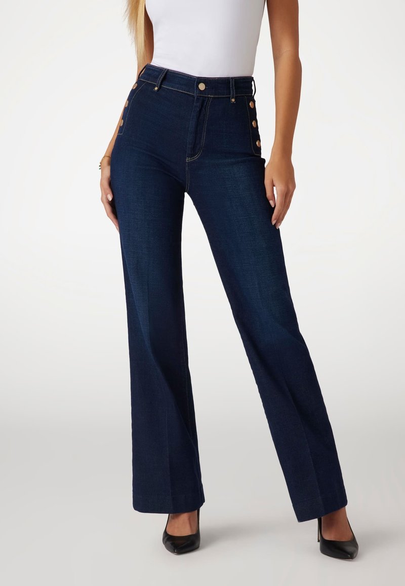 Högmidjade mörkblå jeans i denim med utsvängda ben, gyllene knappdetaljer på sidan och standard dragkedjestängning. Slät textur, välarbetad passform.