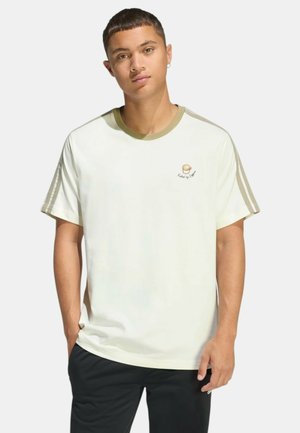 Giovane uomo con capelli corti che indossa una maglietta bianca con strisce beige sulle maniche e una piccola grafica rotonda sul petto, pantaloni neri, in piedi.