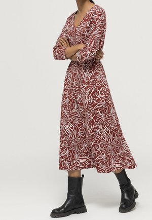 Robe midi rouge avec un motif floral abstrait blanc, décolleté en V, manches longues et taille froncée. Associée à des bottines noires.