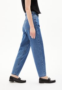 Ljusblå höga raka jeans med fransar i fållen, kombinerat med svarta loafers med vita detaljer, visade från sidan.