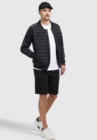Giubbotto imbottito nero con un'estremità in tessuto liscio, t-shirt bianca, pantaloni corti neri e sneaker bianche con accenti neri; modello che indossa un cappellino.