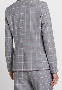 Osoba w szarym garniturze w kratę, z widokiem z tyłu, na jednolitym jasnym tle.