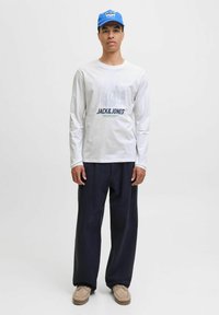 Jonge man in een blauwe pet, een witte lange mouwen grafische shirt, losse donkere broek en beige moccasinstijl schoenen staat tegen een effen achtergrond.