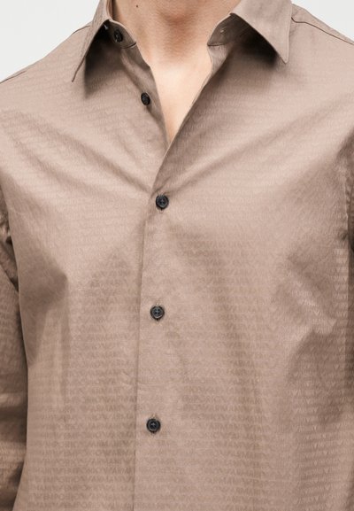 Μπεζ μακρυμάνικο πουκάμισο με διακριτικό μοτίβο λογότυπου Armani και μαύρα κουμπιά, φορεμένο κουμπωμένο εκτός από τα δύο πάνω κουμπιά που είναι ανοικτά.