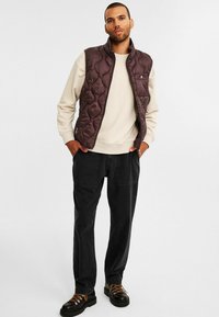 Mand iført en mørkebrun quilted vest over en beige sweatshirt, sorte bukser og sorte støvler med orange snørebånd, står med hænderne i lommerne.