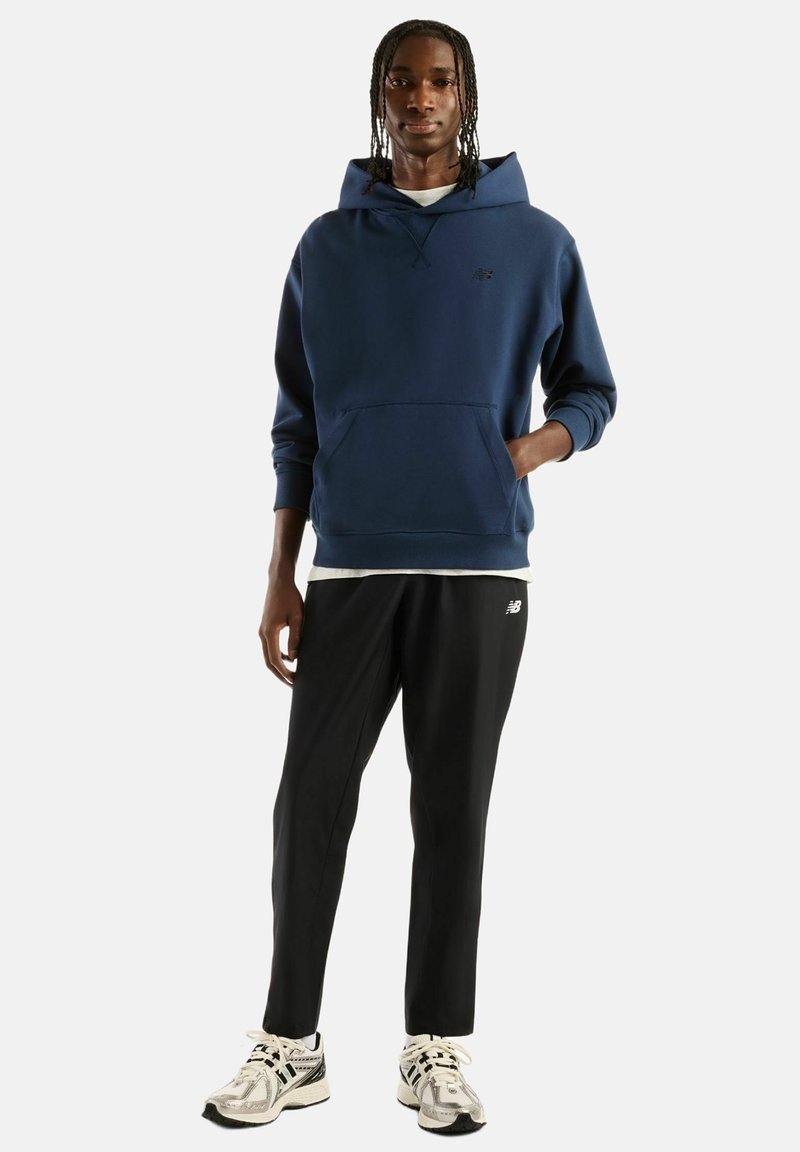 Young man with braided hair wearing a navy hoodie, black pants, and white sneakers standing against a plain background.