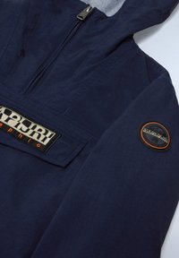 Marineblå lett jakke med glidelås foran, teksturert stoff og brodert logo-patch på brystet og ermet.