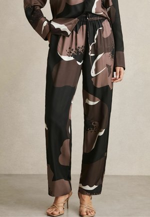 Femme portant un pantalon large noir et marron avec un grand imprimé floral, associé à un haut à manches longues assorti et des sandales beige à talons hauts.