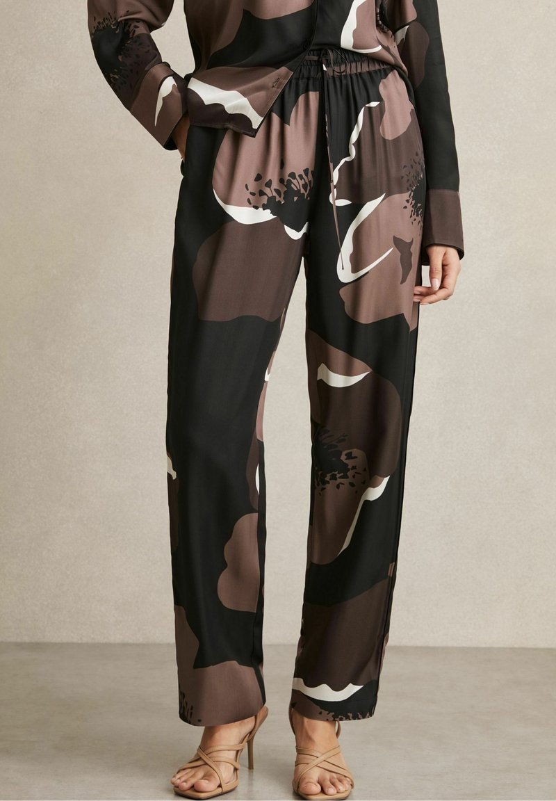Femme portant un pantalon large noir et marron avec un grand imprimé floral, associé à un haut à manches longues assorti et des sandales beige à talons hauts.