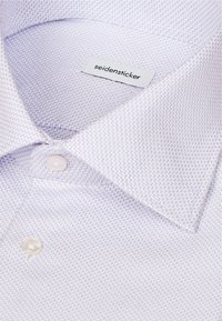 Gros plan d'une chemise blanche avec un motif zigzag bleu fin, des boutons visibles, et une étiquette de marque Seidensticker à l'intérieur du col.