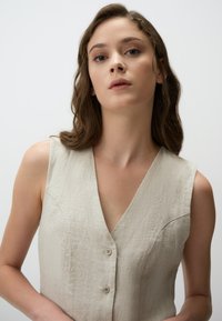 Débardeur en lin sans manches en beige clair avec un col en V, présentant une fermeture à boutons et une texture douce, une coupe ajustée et des détails de couture discrets.