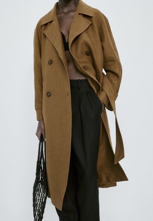 Personne portant un manteau marron croisé, un pantalon noir taille haute, tenant un sac fourre-tout en filet noir, une main dans la poche.