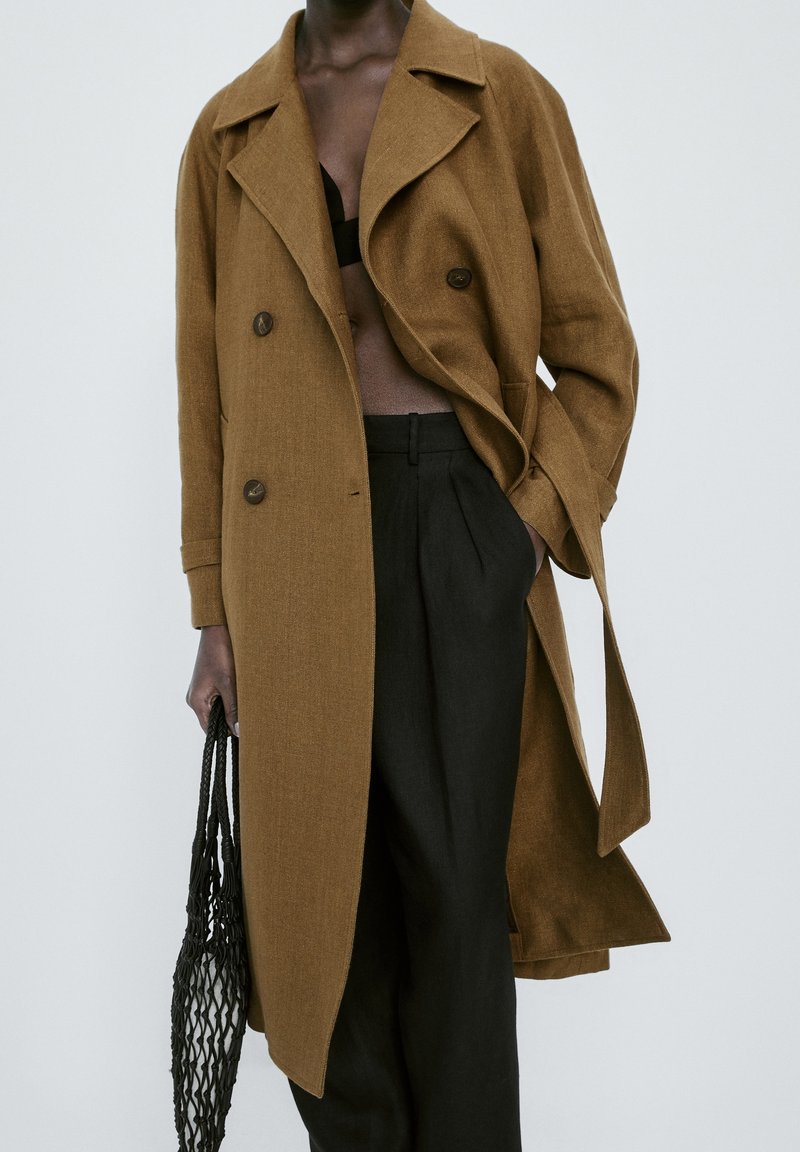 Personne portant un manteau marron croisé, un pantalon noir taille haute, tenant un sac fourre-tout en filet noir, une main dans la poche.