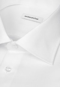 Gros plan sur le col d'une chemise blanche avec une étiquette affichant le nom de la marque "seidensticker" et un bouton blanc visible.