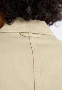 Beige jacket with a smooth texture, featuring a wide collar, back seam detail, and clean stitching along the edges.