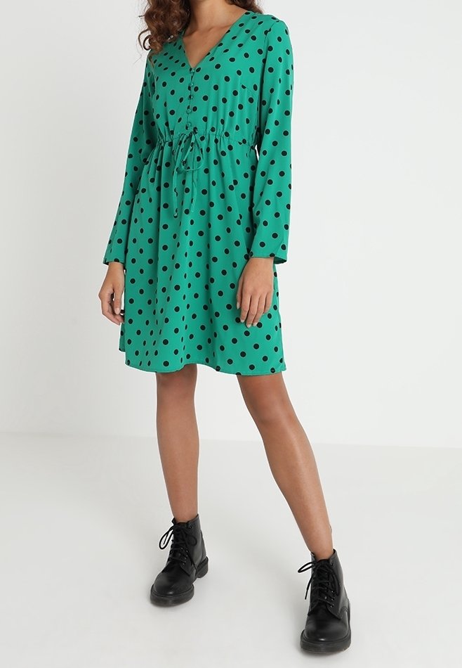 Femme portant une robe verte à manches longues avec des pois noirs et des bottines noires à lacets, debout sur un fond blanc uni.