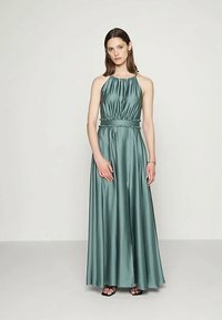 Mulher em pé, vestindo um vestido maxi de cetim teal sem mangas, com um decote halter e cintura ajustada, e sandálias pretas de tiras.