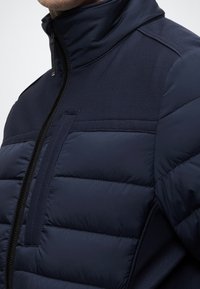 Tengerészkék puffdzseki magas gallérral, steppelt textúrával és hálós panelekkel. Cipzáras elülső rész kontrasztos varrással és hurok részlettel.