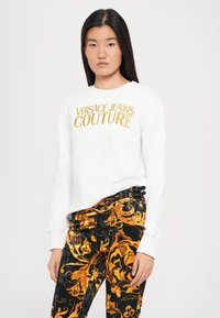 Felpa bianca con testo dorato "VERSACE JEANS COUTURE", abbinata a pantaloni neri in velluto con un intricato motivo barocco dorato.