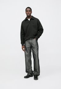 Melnā pulovers ar rāvējslēdzēja apkakli un krūšu logotipu, kombinēts ar tumši pelēnām džinsām un melniem apaviem, attēlots uz neitrāla fona.