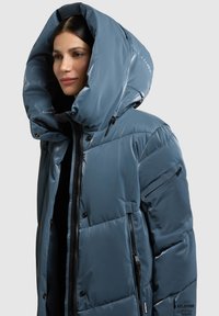 Prošívaná bunda s kapucí v barvě břidlicově modré. Má lesklý povrch, kapsy na zip a vysoký límec. Charakteristické švy panelů a značení.