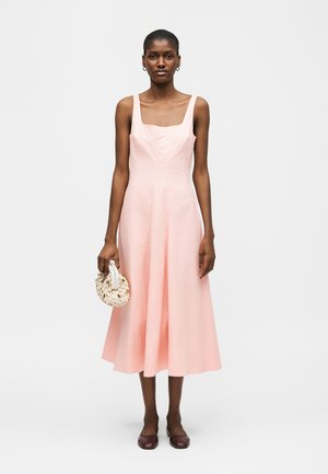 Donna che indossa un vestito midi rosa senza maniche, tiene in mano una borsa bianca intrecciata e indossa scarpe piatte scure, in piedi davanti a uno sfondo semplice.