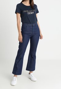 Žena v námořnickém tričku "Marc O'Polo Denim", v tmavých širokých džínách s vysokým pasem a v bílých teniskách, stojící na jednobarevném pozadí.