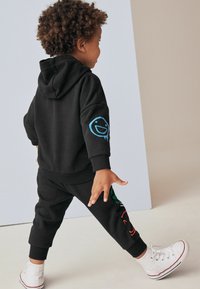 Sort hættetrøje og sweatpants sæt med et blåt smiley-design på armen og farverige grafikker på benet, parret med hvide sneakers.