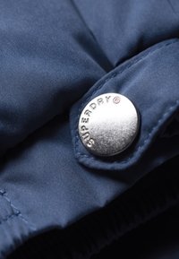 Marineblauwe jas geweven met een gestructureerde afwerking, met een prominente zilveren drukknoop met de inscriptie "SUPERDRY." Gedetailleerde stiksels zichtbaar.