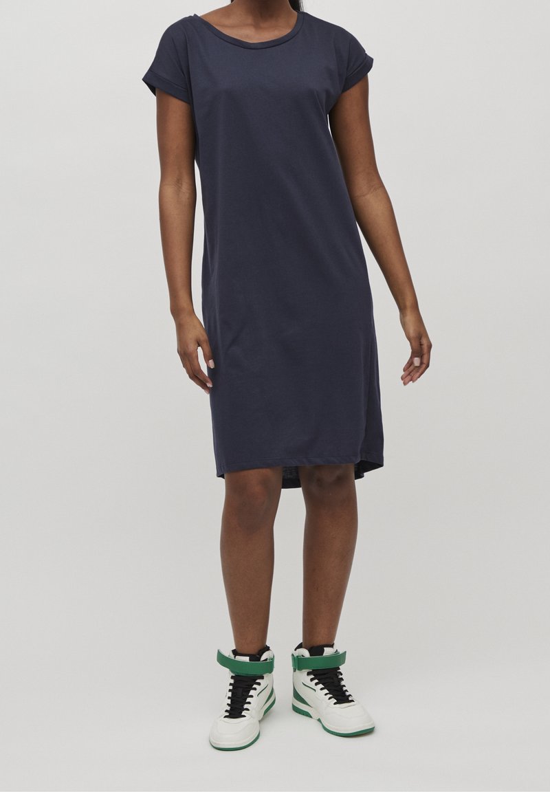 Personne portant une robe bleu marine à manches courtes longueur genou et des baskets montantes blanches avec des détails verts sur un fond uni.