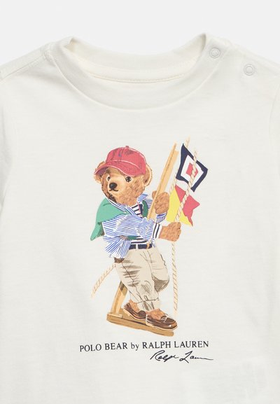 Αρκουδάκι με κόκκινο καπέλο, ριγέ πουκάμισο, μπεζ παντελόνι και μοκασίνια, κάθεται σε ξύλινη κούνια κρατώντας ναυτικές σημαίες.