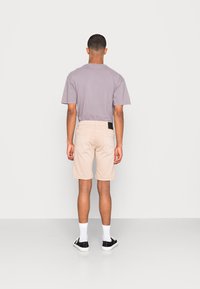 Lyhythihainen laventelit-paita yhdistettynä beigeihin shortseihin, joissa on takataskut ja musta etiketti vyötäröllä. Valkoiset sukat ja mustat kengät.