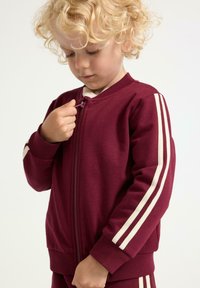 Burgunderfarvet zip-jakke med off-white striber langs skuldrene. Blød, bomuldsblanding; ribbede manchetter og talje; frontlommer.