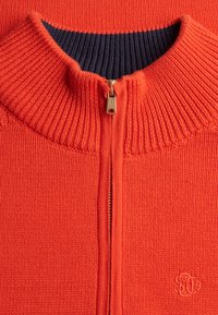 Nahaufnahme eines orangefarbenen Strickpullovers mit geripptem Kragen, goldenem Reißverschluss und sichtbar navyblauem Innenfutter am Halsausschnitt.