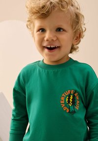 Sudadera de algodón verde con un logo redondo que presenta árboles y la frase "LA AVENTURA TE ESPERA" en naranja y negro. Cuello redondo clásico y mangas largas.