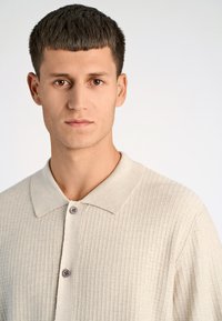 Lichtbeige geribde polo met een platte kraag, voorzien van twee bruiskleurige knopen en een gestructureerde stof, die een schone en minimalistische uitstraling biedt.