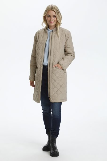 Manteau beige matelassé avec un col haut, des poches avant et une coupe droite. Porté sur une chemise bleu clair, associé à un jean foncé et des bottes noires.
