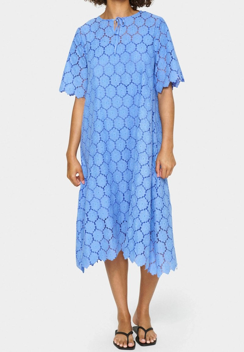 Femme portant une robe en dentelle bleue mi-longue avec des motifs circulaires, des manches courtes, des bords festonnés et un petit col en trou de serrure noué, avec des sandales noires.