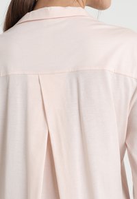 Blusa rosa chiaro con una consistenza morbida, caratterizzata da una piega sul retro e un colletto. Design semplice, che sottolinea il comfort casual.