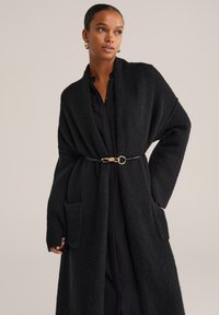 Sieviete ar garu melnu teksturētu kardigānu ar lielām kabatām, sašaurinātu jostasvietā ar plānu melnu jostu ar zelta sprādzi.