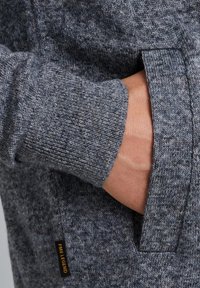 Gray knit sweater with a textured pattern, featuring a pocket on the side and a visible brand label. Soft material and ribbed cuffs.
