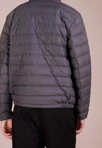 Grå pufferjacka med horisontellt kviltat mönster, elastisk nederkant och långärmar, sedd bakifrån med en slät yta.