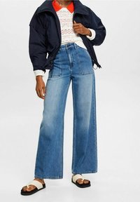 Hochtaillierte, weit geschnittene blaue Jeans mit genähten Taschen und einem rohen Saum, kombiniert mit einer marineblauen Jacke und weißen Sandalen mit offenen Zehen.