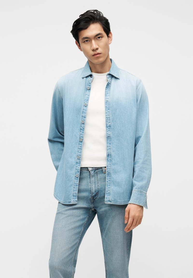 Mann trägt ein hellblaues Jeanshemd offen über einem weißen T-Shirt und hellblaue Jeans, steht vor einem schlichten hellen Hintergrund.