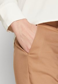 Main partiellement dans la poche avant d'un pantalon marron clair porté avec une chemise crème à manches longues.