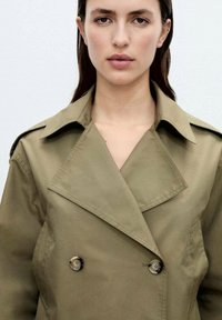 Olive green trench coat with a double-breasted front, large notched collar, and smooth texture; features round buttons and side pockets.