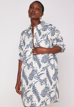 Donna di colore che indossa un abito camicia bianco con stampe di pavone blu e fogliame, maniche arrotolate e orecchini dorati, in posa su uno sfondo neutro.